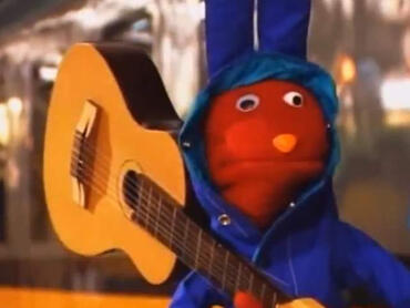 A pic of Bodoque from 31 minutos. He is a red rabbit puppet, he is wearing a raincoat and is holding a guitar over his shoulder